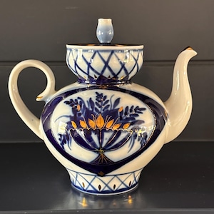 May include: A white porcelain teapot with a blue and gold floral design. The teapot features a rounded body, a curved spout, and a loop handle. The lid is topped with a small, blue knob. The teapot is decorated with a blue and gold floral pattern.