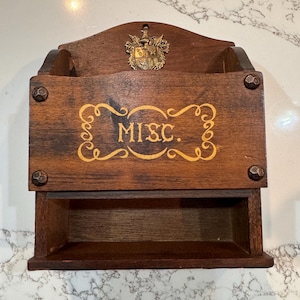 May include: A vintage wooden wall organizer with a dark brown finish. The organizer features a top shelf, a main compartment with the word "MISC." in gold lettering, and a bottom drawer. Decorative metal accents and a crest add detail.