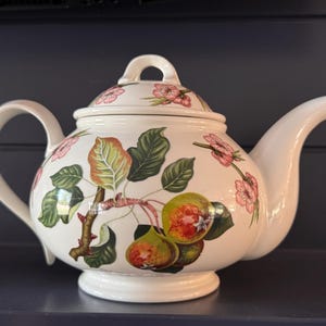 May include: A white ceramic teapot featuring a botanical illustration of green leaves, brown branches, and pink blossoms. The design includes two round green and yellow fruits on the front. The teapot has a matching lid with a handle.
