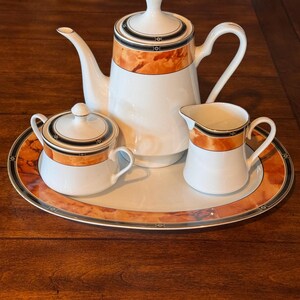 May include: A white porcelain tea set consisting of a teapot with a lid, a lidded sugar bowl, a creamer, and an oval serving tray. Each piece features a decorative band with a marbled orange pattern, black trim, and thin gold accents.