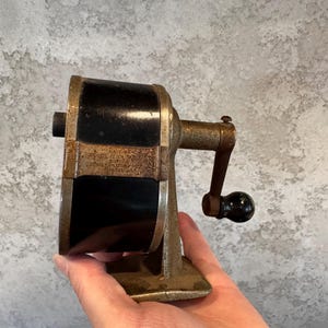 May include: A vintage manual pencil sharpener featuring a black cylindrical body with a weathered, textured metal frame and base. It includes a side-mounted hand crank with a small black knob, showing signs of age and oxidation on the metal parts.
