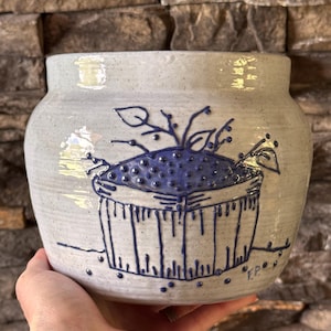 May include: A handmade ceramic pot with a light gray speckled finish. The pot features a blue illustration of a basket filled with berries and leaves. The pot is held in a person's hand.