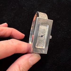 May include: A silver Gucci watch with a rectangular face and a textured band. The watch face displays the Gucci logo and the words "Gucci Italy". The watch is held against a black background.