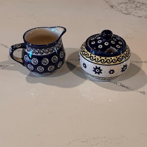 May include: A ceramic creamer pitcher and a matching lidded sugar bowl. Both pieces feature a white base with hand-painted blue and yellow patterns, including swirls, dots, and star-like floral motifs. The creamer has a handle and spout.