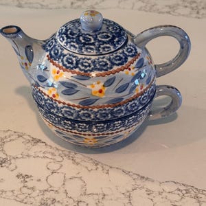 May include: A blue and white ceramic teapot with a matching teacup. The teapot has a rounded shape with a lid and a handle. The design features floral and geometric patterns in blue, yellow, and brown. The teacup is stacked beneath the teapot.