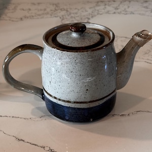 May include: A ceramic teapot with a speckled white and gray body, a dark blue base, and a brown lid. The teapot has a curved handle and a spout. The lid has a small knob for easy removal.