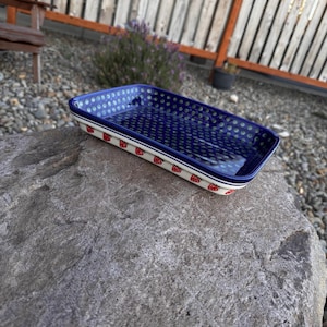 May include: A rectangular ceramic baking dish featuring a deep blue interior with a pattern of small white dots. The exterior sides are white and decorated with a repeating pattern of small red strawberries and green leaves along the rim.