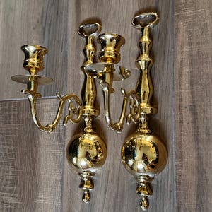 May include: A pair of polished gold-tone metal wall sconces, each featuring a single candle holder arm extending from a vertical stem. The design includes a prominent spherical base, a slender neck, and a top loop for mounting on a wall surface.