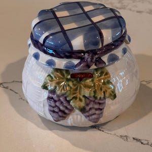 May include: A white ceramic jar with a rounded body featuring a textured basket-weave pattern. The lid is painted with a blue and white plaid design and a blue polka-dot rim. Two clusters of purple grapes with green leaves are embossed on the front.