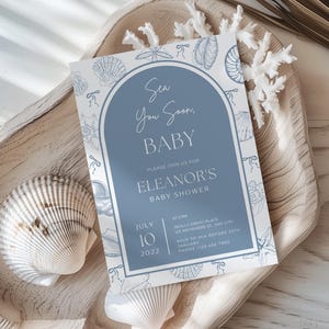 May include: A baby shower invitation with a blue arch design and the text "Sea You Soon, BABY". The invitation is surrounded by seashell illustrations and the details for Eleanor's baby shower. The invitation is on a wooden tray with seashells and coral.