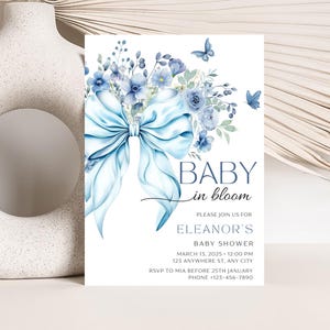 May include: Blue and white baby shower invitation with a watercolor illustration of a blue bow and flowers. The text reads "Baby in Bloom" and includes the details for Eleanor's baby shower.