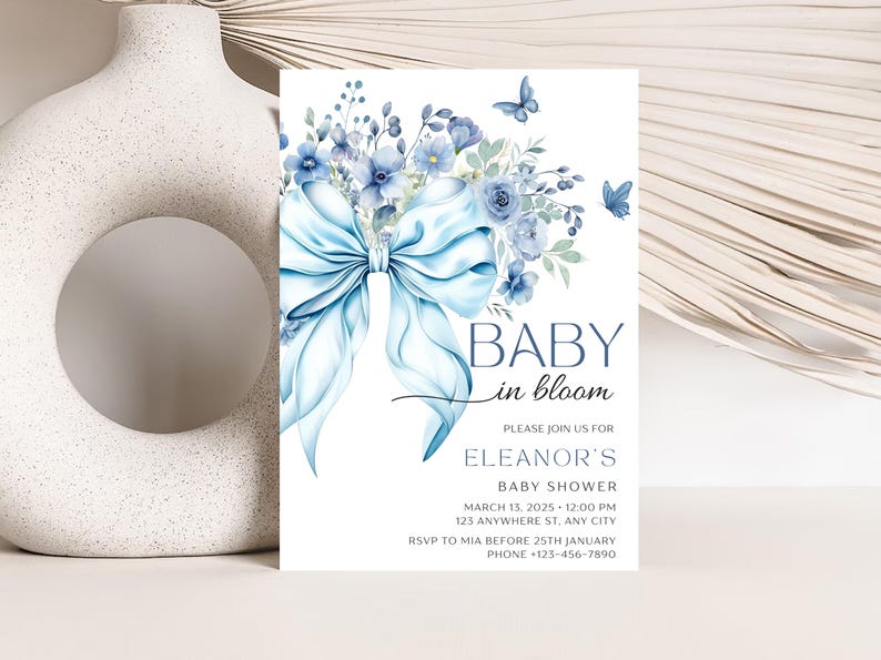 May include: Blue and white baby shower invitation with a watercolor illustration of a blue bow and flowers. The text reads "Baby in Bloom" and includes the details for Eleanor's baby shower.