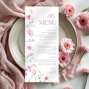 May include: A floral menu card with pink flowers and butterflies, with the text "The Menu". The menu lists appetizers, main courses, desserts, and drinks. The card is on a plate with pink flowers and fabric. The overall theme is a wedding or special event.