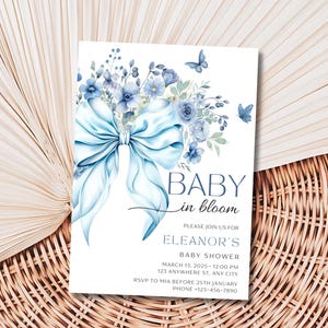 May include: Blue and white baby shower invitation with a watercolor illustration of a blue bow and flowers. The text reads "Baby in Bloom" and "Eleanor's Baby Shower".