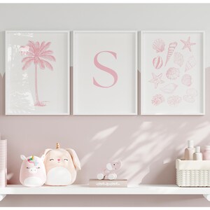 Könnte beinhalten: Drei gerahmte Drucke mit rosa Illustrationen einer Palme, dem Buchstaben "S" und Muscheln. Unterhalb befindet sich ein weißes Regal mit Plüschtieren, Büchern und Dekorationsartikeln. Das Farbschema ist zartrosa und weiß.