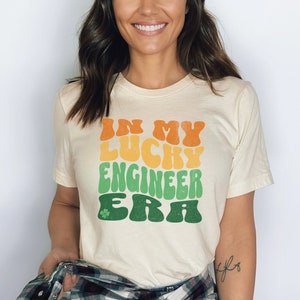 May include: Cream-colored t-shirt with the text "IN MY LUCKY ENGINEER ERA" in orange, yellow, and green, with a green shamrock. The shirt has short sleeves and a crew neck.