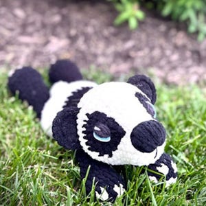 May include: A handmade crochet panda bear plush toy in black and white, lying on green grass. The panda has black ears, eyes, and paws, with a white face and body. The toy is soft and cuddly, perfect for children.