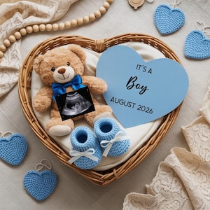 Pode incluir: Um ursinho com laço azul, imagem de ultrassom e sapatinhos azuis estão em uma cesta de vime em forma de coração. Uma placa azul em forma de coração diz "It's a Boy August 2026". Corações azuis de crochê estão espalhados.