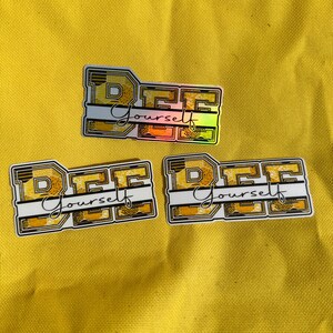 Könnte beinhalten: Drei goldene und schwarze Aufkleber mit den Worten "Bee Yourself" in weißen Buchstaben. Die Aufkleber haben ein Bienenwabenmuster.