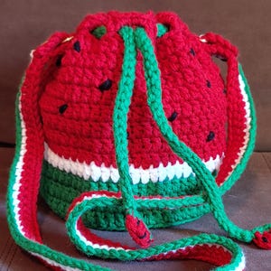 Könnte beinhalten: Eine gehäkelte Wassermelonen-Kordeltasche mit einem grün-weiß-roten Band. Die Tasche ist rot mit schwarzen Kernen und hat einen grün-weißen Streifen am Boden.