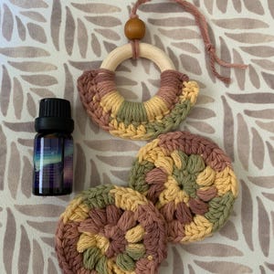 May include: An essential oil bottle and a set of handmade crochet items in shades of yellow, green, and mauve. The set includes a wooden ring with crochet detail and two round coasters. The oil bottle has a black cap.