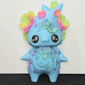May include: A light blue figurine with a smooth matte finish, featuring large black eyes with white highlights. The creature is adorned with pastel-colored floral accents in shades of pink, yellow, and green on its head and chest area.