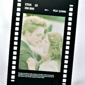 May include: A black card in the shape of a film strip, displaying a romantic image of a couple. The card has white text at the top and bottom, including "STAX 15 ISO 800" and "F5.6 1/1000". A purple paper clip secures the card.