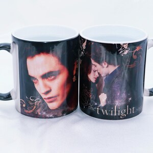 May include: Two ceramic mugs with black handles and rims. Each mug features a different image of characters from the Twilight saga. The text "the twilight saga" is visible on one mug.