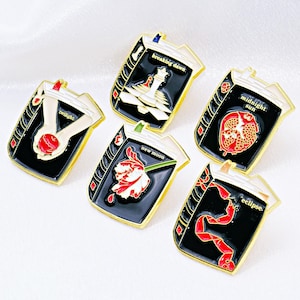 Może przedstawiać: Pięć emaliowanych przypinek w kształcie książek ze złotymi obramowaniami. Każda przypinka przedstawia inny projekt okładki i tytuł: "Twilight", "Breaking Dawn", "Midnight Sun", "New Moon" i "Eclipse". Przypinki mają czarne tła i kolorowe ilustracje.