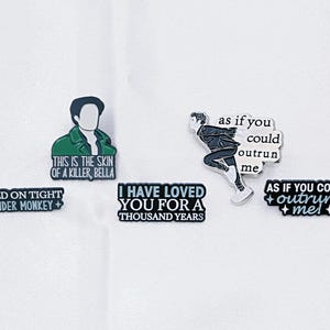 Puede incluir: Un conjunto de pines esmaltados con varios diseños y texto. Un pin presenta una figura con el texto "THIS IS THE SKIN OF A KILLER, BELLA." Otros pines incluyen frases como "HOLD ON TIGHT + SPIDER MONKEY +" e "I HAVE LOVED YOU FOR A THOUSAND YEARS."