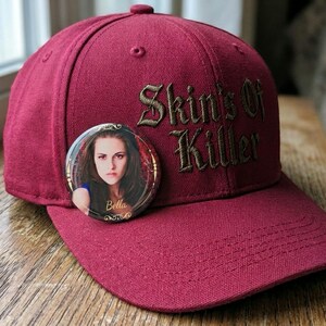 Peut inclure: Une casquette de baseball bordeaux avec le texte brodé "Skin's Of Killer" en fil foncé. Un badge rond est fixé sur le panneau avant, montrant un portrait photographique d'une personne aux cheveux bruns avec le nom "Bella" inscrit.