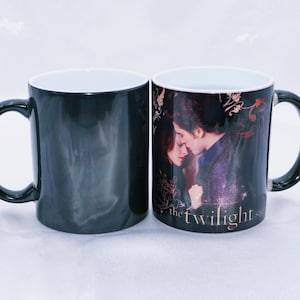 Może przedstawiać: Dwa czarne ceramiczne kubki. Jeden kubek jest w jednolitym czarnym kolorze, drugi przedstawia romantyczny obraz dwóch osób z napisem "the twilight saga". Oba kubki mają białe wnętrze i czarny uchwyt.