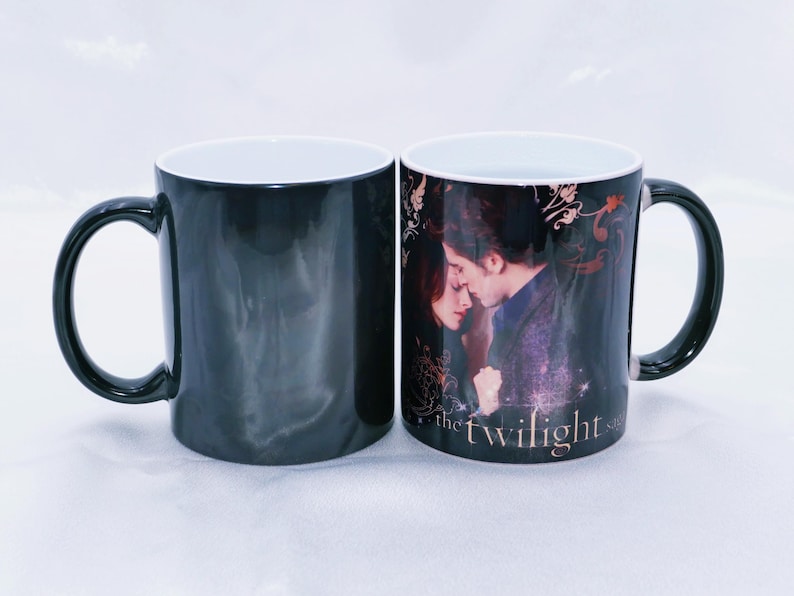 May include: Two black ceramic mugs. One mug is solid black, the other features a romantic image of two people with the text "the twilight saga". Both mugs have a white interior and black handle.