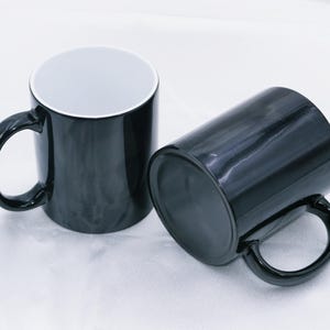 May include: Two black ceramic mugs with a glossy finish. One mug is upright, showcasing a white interior, while the other lies on its side. Both mugs have a rounded handle and are set against a white background.