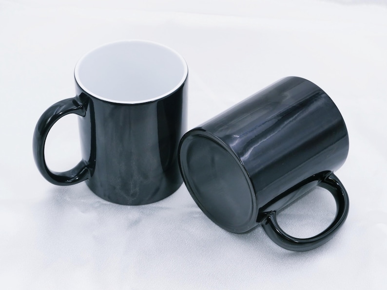 May include: Two black ceramic mugs with a glossy finish. One mug is upright, showcasing a white interior, while the other lies on its side. Both mugs have a rounded handle and are set against a white background.
