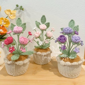 May include: Three crochet flower arrangements in beige pots. The flowers are pink, purple, and white, with green leaves. The pots have a scalloped edge. The text "Tinycute Studio" is on the front of the pots.