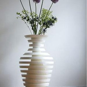 Könnte beinhalten: Eine weiße Keramikvase mit einem einzigartigen, geschichteten Design, die einen Strauß lilafarbener Allium-Blumen hält.