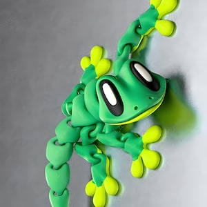 May include: A green and yellow articulated toy frog with large eyes and a long tail. The frog is posed as if it is climbing a wall.