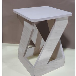 May include: A white wooden stool with a simple, geometric design. The stool has a square top and a base that resembles a stylized 'Z' shape.