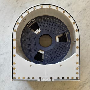 Può includere: Supporto per bobina di filamento per stampante 3D bianco e blu navy con la scritta "PRISM" stampata sul davanti. Il supporto ha un disco perforato blu navy circolare all'interno, che contiene filamento bianco. Il supporto è rettangolare con bordi arrotondati.