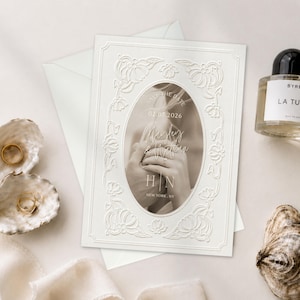Könnte beinhalten: Eine weiße, rechteckige Save-the-Date-Karte mit geprägtem Blumenrand und einem zentralen ovalen Foto von Händen mit Ringen. Der Text lautet: Save the Date, 02.07.2026, Namen, H | N und New York, NY. Begleitet von zwei Austernschalen und einem Parfümflakon.
