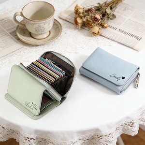 May include: Two small leather wallets, one mint green and one light blue, are on a white surface. The mint green wallet is open, showing multiple credit cards. The blue wallet is closed and has the name "Emily" printed on it. Both wallets have zippers.