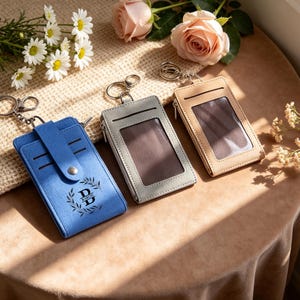 Peut inclure: Trois porte-cartes rectangulaires avec anneaux porte-clés, comprenant un modèle bleu avec une patte à pression et la lettre B avec le texte BARON, un modèle gris et un modèle beige. Chaque porte-cartes possède une fenêtre transparente.
