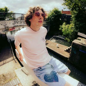 Pode incluir: Uma pessoa jovem vestindo uma camiseta rosa com um pequeno design gráfico e shorts de jeans azul com um remendo em forma de coração. A pessoa está sentada em uma superfície branca perto de um canal.