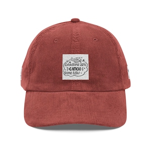 May include: A rust-colored corduroy baseball cap with a curved brim. The cap features a square patch with the text "Catch Some Zz's, Catch Some Fish!" and a fish illustration. The hat has a classic six-panel design.