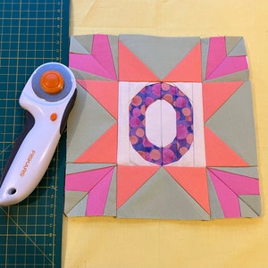 May include: A colorful quilt block with a central letter "O" design, surrounded by geometric shapes in pink, coral, and green. A white and orange rotary cutter is positioned on a green cutting mat, next to the quilt block. The background is yellow.