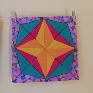 May include: A colorful, geometric quilt block with a starburst design. The center features a yellow and orange star shape, surrounded by pink and teal triangles. The border is a blue fabric with pink and orange polka dots. The quilt block is hanging on a clothesline.