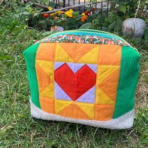 May include: A colorful, quilted pouch with a heart design. The pouch features a red heart in the center, surrounded by orange, yellow, and purple geometric shapes. The sides are green corduroy, and the top has a strawberry pattern. The base is white, fluffy material.