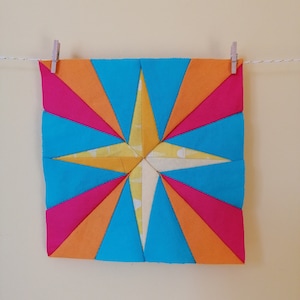 May include: A colourful patchwork quilt block featuring a star design in shades of blue, orange, and pink. The star is made from multiple fabric pieces sewn together.