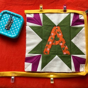 May include: A quilt block with a green star and the letter "A" in orange fabric with a floral print. The block is surrounded by a yellow fabric border and is pinned to a red surface with clips.